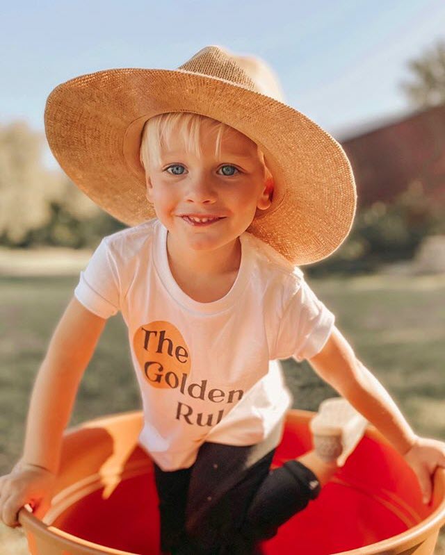 Golden Rule tee shirt smiling toddler boy wearing a large straw hat, standing in a flower pot with a shirt printed with the words Golden Rule